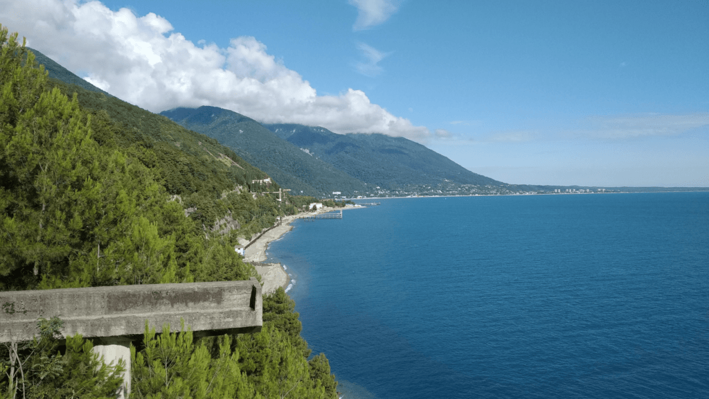 Coastal landscape in Abkhazia featuring deep blue sea, lush green mountains, and a clear sky. A road winds between the forest and the shoreline, with a distant view of a seaside city.