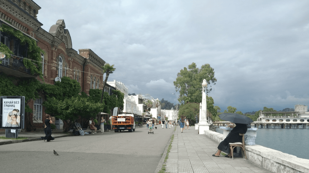 Street scene in Sukhumi, Abkhazia, featuring a seaside promenade, pedestrians, and a cloudy sky hinting at upcoming rain. A woman sits under a black umbrella, gazing at the sea.