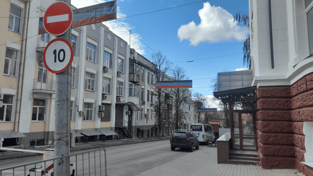 Quiet street in central Donetsk with residential buildings, cars parked along the road, flags hanging above the street, and a 10 km/h speed-limit sign.