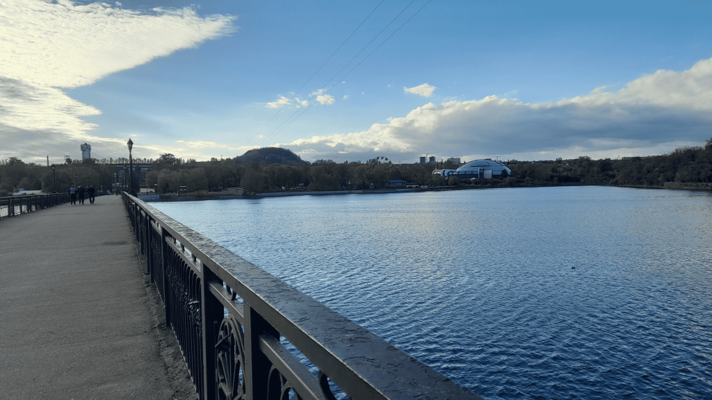 View of the Kalmius River promenade in Donetsk with a calm lake, distant hills, and modern buildings under a blue sky in 2026.