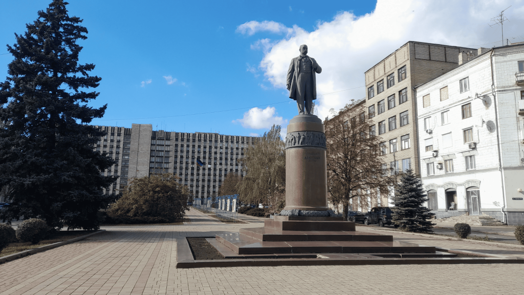 Monument on a raised pedestal in Donetsk’s Shevchenko Square, with surrounding buildings and a clear blue sky.