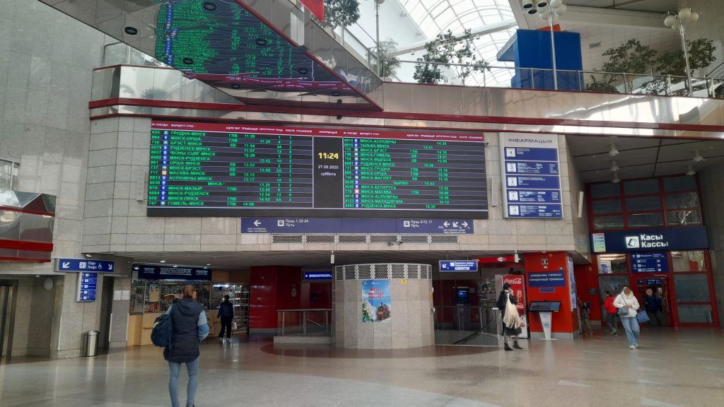 Departure boards and main hall inside Minsk railway station, with travelers and ticket offices.